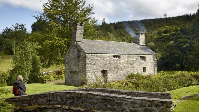 Ymwelydd yn eistedd ar bont fwa fach, yng nghefn gwlad yn Nhŷ Mawr Wybrnant, Conwy. Mae’n edrych tuag at adeilad carreg sydd â mwg yn dod allan o’r simnai.